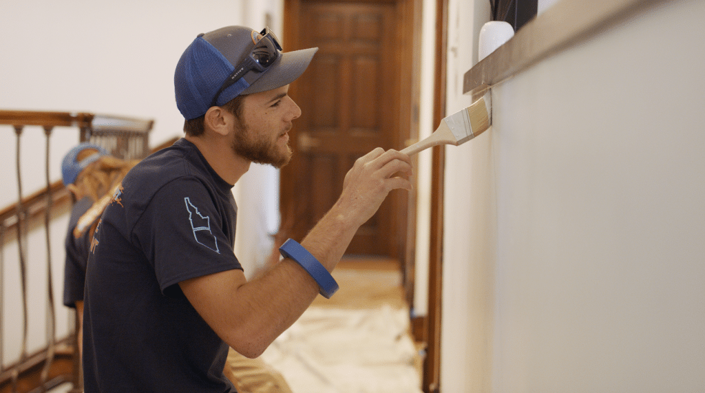 man with beard painting interior of a house with white paint, wearing a cap and sunglasses on top