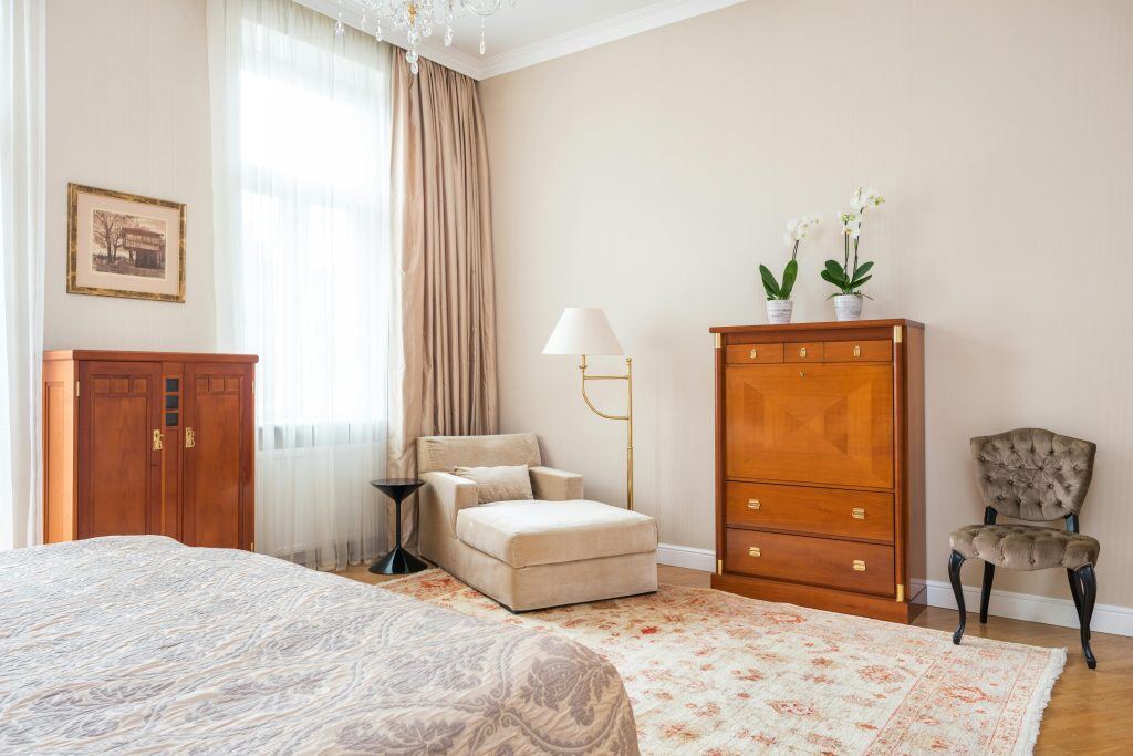 Bedroom with beige walls. 2 chairs are against the wall, with 2 cabinets next to them. There is a lamp, decorative flowers and a frame.